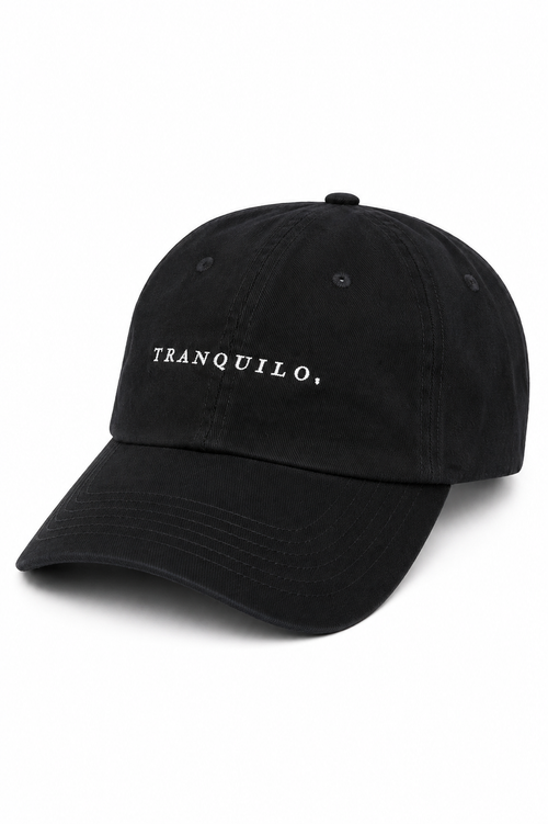 Boné preto de aba curva, fotografado em fundo claro, com bordado frontal branco escrito “TRANQUILO,” em letras maiúsculas. A peça tem costuras aparentes nos painéis, ilhoses no topo e acabamento em tecido preto.