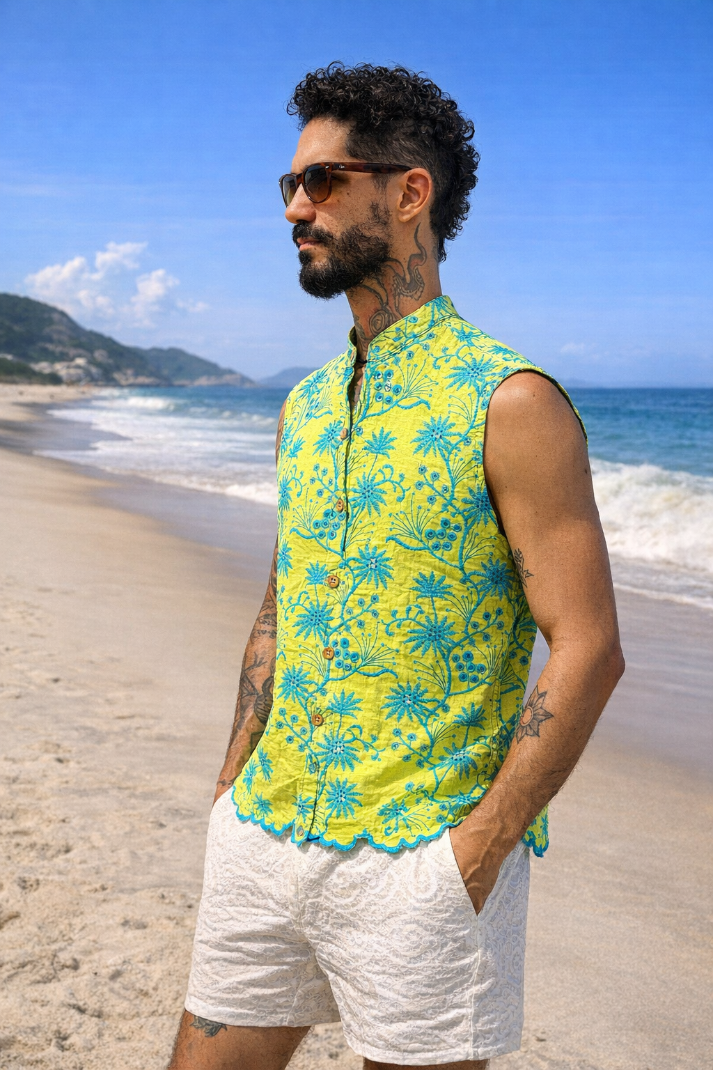 Pessoa usando camisa sem manga verde-lima com estampa azul-turquesa, gola padre e botões de coco em uma praia.