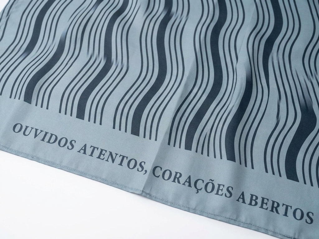 Foto tratada em close da Canga Ondas Tranquilo, mostrando textura do tecido cinza-azulado, linhas onduladas escuras e trecho da faixa textual “OUVIDOS ATENTOS, CORAÇÕES ABERTOS”.