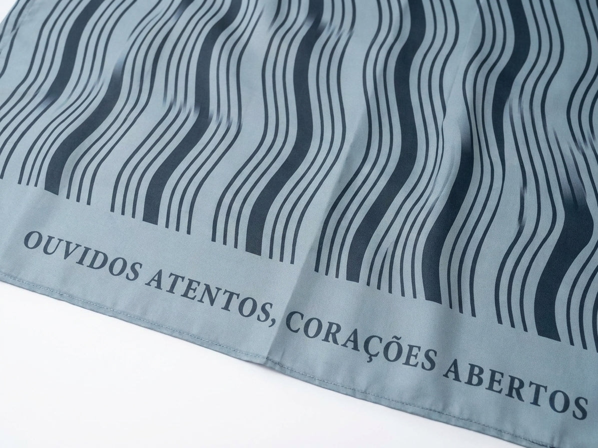 Foto tratada em close da Canga Ondas Tranquilo, mostrando textura do tecido cinza-azulado, linhas onduladas escuras e trecho da faixa textual “OUVIDOS ATENTOS, CORAÇÕES ABERTOS”.