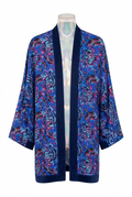 Kimona aberta em tecido azul intenso com estampa floral em tons de azul, rosa, branco e preto. A peça tem mangas amplas, comprimento alongado, pala frontal e barras em azul-marinho, com etiqueta oRnii visível na parte interna da gola.