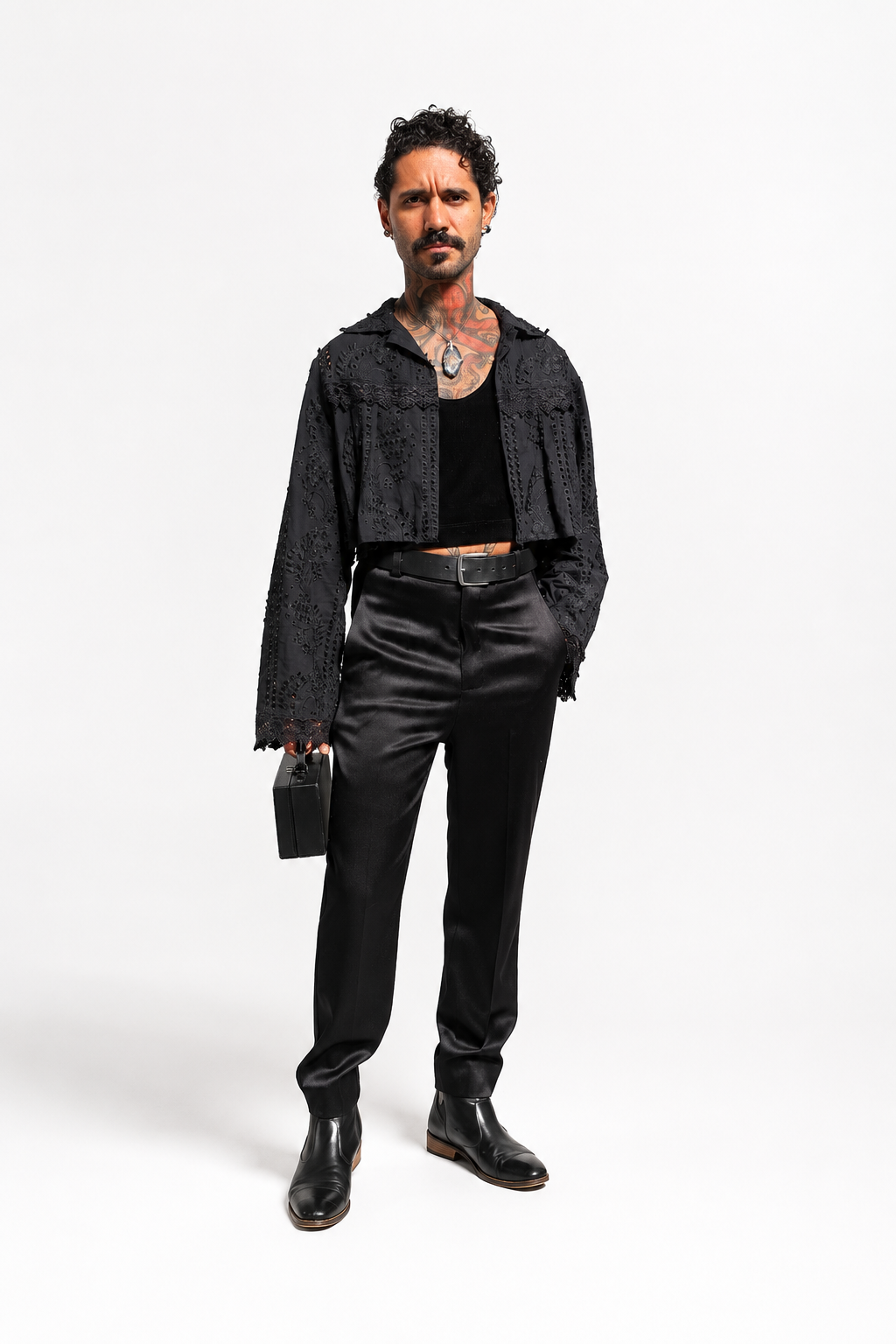 Modelo em pé sobre fundo claro usando o Bolero Morcego preto aberto sobre top preto, calça preta de brilho acetinado, cinto preto, botas pretas e bolsa pequena preta. O bolero é curto, tem gola, mangas longas amplas, lese vazada com desenhos de galhos, folhas e flores em relevo e renda guipir aplicada nas mangas.