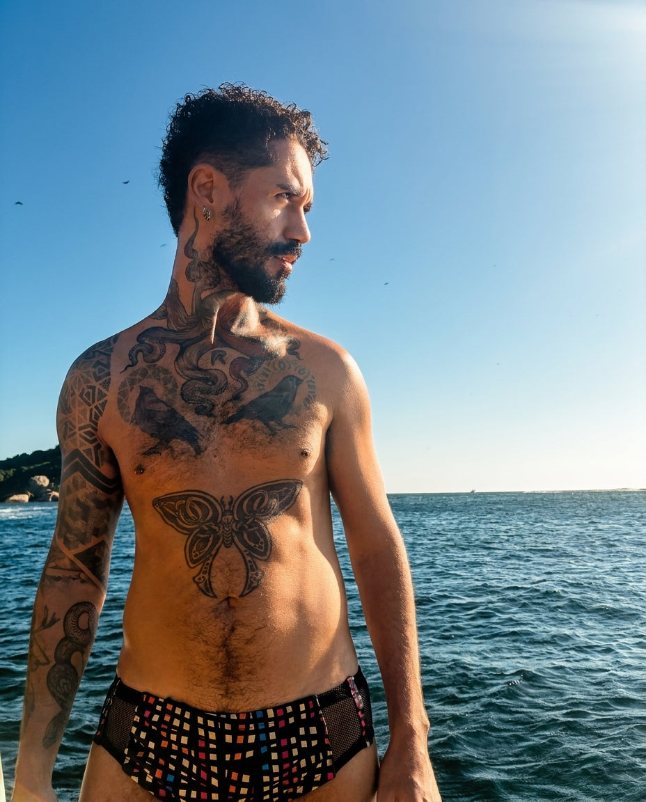 Rafinha Medeiros à beira-mar, com céu azul e oceano ao fundo, usando somente a Sunga Píxel em uma fotografia lifestyle vertical.